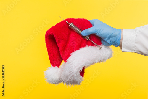 Close-up profile side view of a human hand in blue surgical gloves, holding a Santa Claus hat and a protective mask against COVID-19 coronavirus infections and an antibiotic syringe,