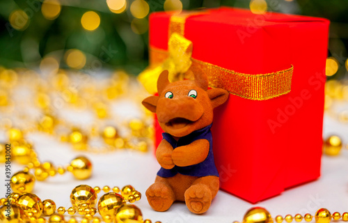 Toy brown bull on a red gift box on a white background. Christmas tree branch with red toys and golden garlands in the background. Bull as a symbol of the new year 2021. New Year, Christmas concept.