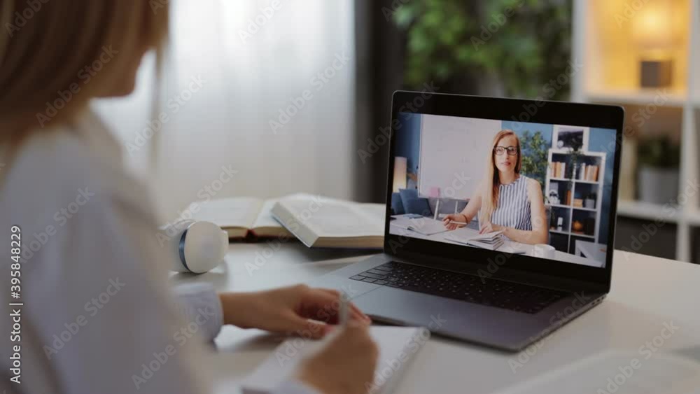 Back view of young woman watching online lesson on wireless laptop while staying at home. Female student taking notes during distance studying. Back view of young woman watching online lesson on wireless laptop while staying at home. Female student taking notes during distance studying.