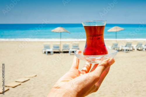 Small glass cup with tea on the background of the blue sea,  beach and beach umbrellas. Hand holds a cup with traditional Turkish tea. 