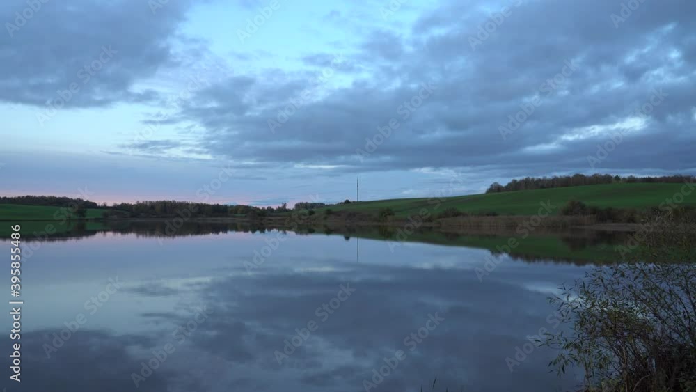 Evening on the lake. A picturesque rural environment in a panoramic scene with a fishpond and farmland. The calm water surface reflects a blue hue cloudy sky in the late afternoon of autumn. Evening on the lake. A picturesque rural environment in a panoramic scene with a fishpond and farmland. The calm water surface reflects a blue hue cloudy sky in the late afternoon of autumn.