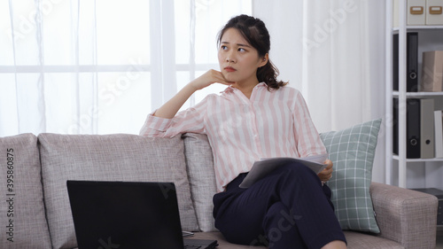 confident asian female ceo with paper and laptop looking outside while working at home office on sofa