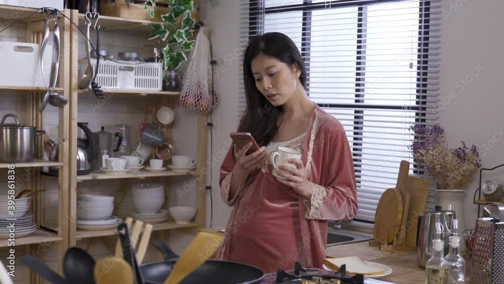 asian woman in slumber wear is reading online news on mobile phone while standing and having breakfast in the kitchen. asian woman in slumber wear is reading online news on mobile phone while standing and having breakfast in the kitchen.