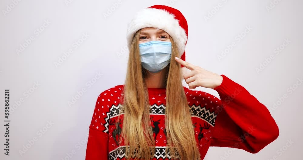 happy young santa claus woman in christmas hat pointing with index finger to medical face mask protected from covid-19 coronavirus virus, on white background, new year celebration holiday happy young santa claus woman in christmas hat pointing with index finger to medical face mask protected from covid-19 coronavirus virus, on white background, new year celebration holiday