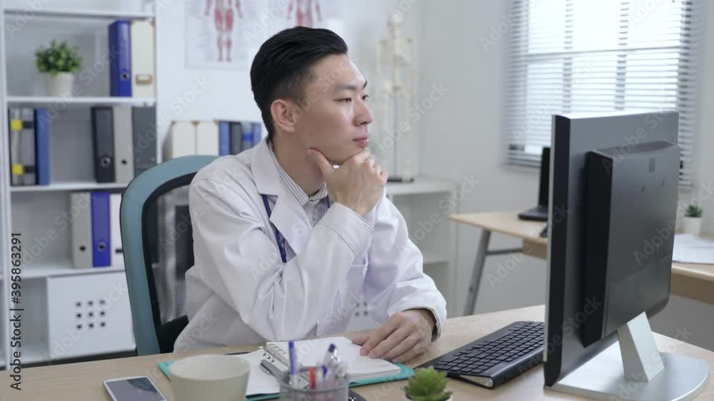 asian specialist looking at the monitor is putting down the pen and touching his chin while making a diagnosis via a video call in his office. asian specialist looking at the monitor is putting down the pen and touching his chin while making a diagnosis via a video call in his office.