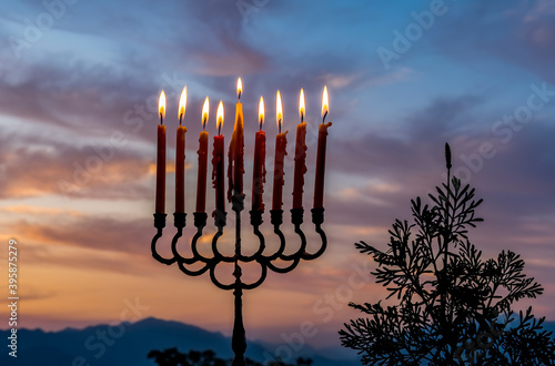 Glitter lights of candles on menorah are traditional symbols for Jewish Hanukkah Holiday of Light. Selective focus on candles. Background with blurred dramatic morning sky and mountains 