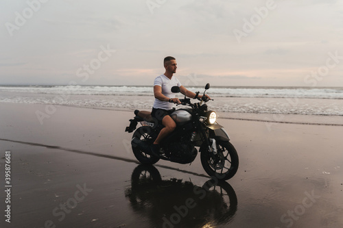 Surfer rides on motorbike with surfboard at sunset ocean beach. Bali island, Indonesia