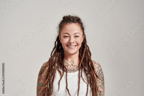 Portrait of young tattooed caucasian woman with dreadlocks wearing white shirt, smiling at camera while posing isolated over grey background