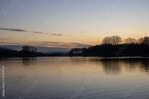 View at sunset with blue sky with some clouds at the horizon reflected on a lake in a cold autumn day  intense orange and dark colors at dusk in Stevenage  Hertfordshire  United Kingdom