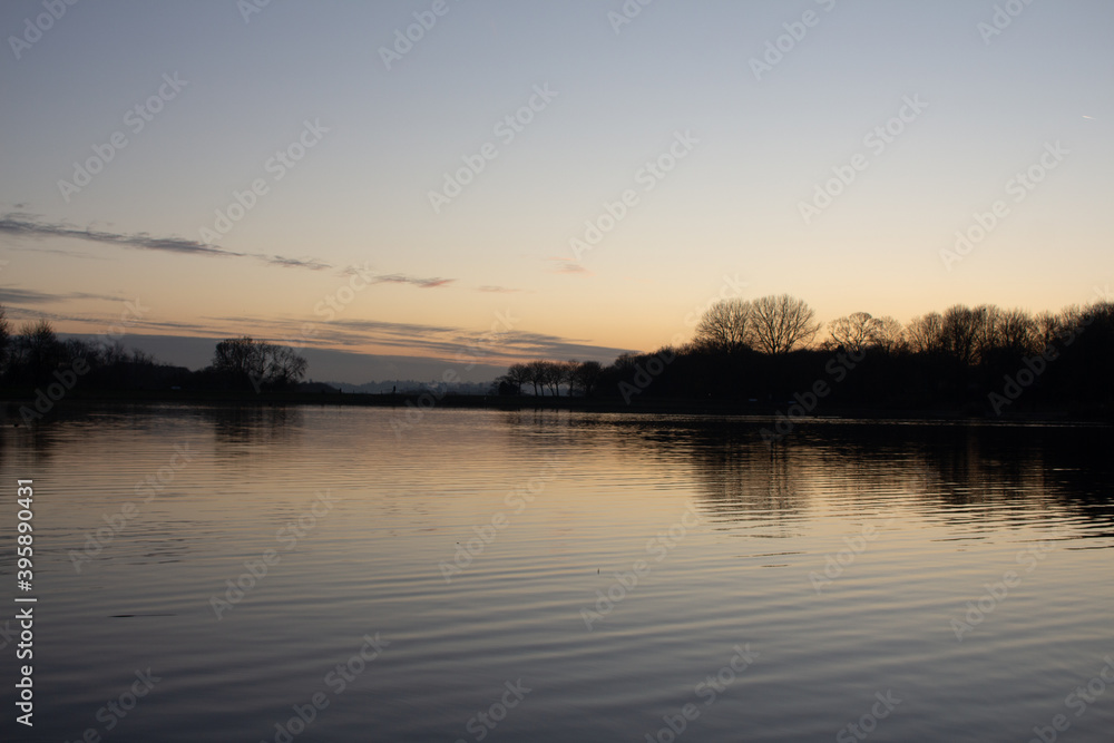 View at sunset with blue sky with some clouds at the horizon reflected on a lake in a cold autumn day, intense orange and dark colors at dusk in Stevenage, Hertfordshire, United Kingdom