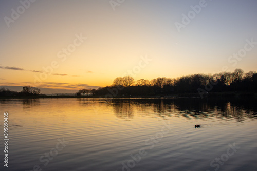View at sunset with blue sky with some clouds at the horizon reflected on a lake in a cold autumn day, intense orange and dark colors at dusk in Stevenage, Hertfordshire, United Kingdom