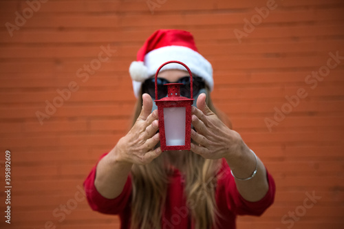 Woman holds in hands red Christmas light. Longhair blond caucasian girl in red sport wear, Christmas hat, sun glasses protective mask. one hand up, wait for present. Orange brick wall behind, copy photo