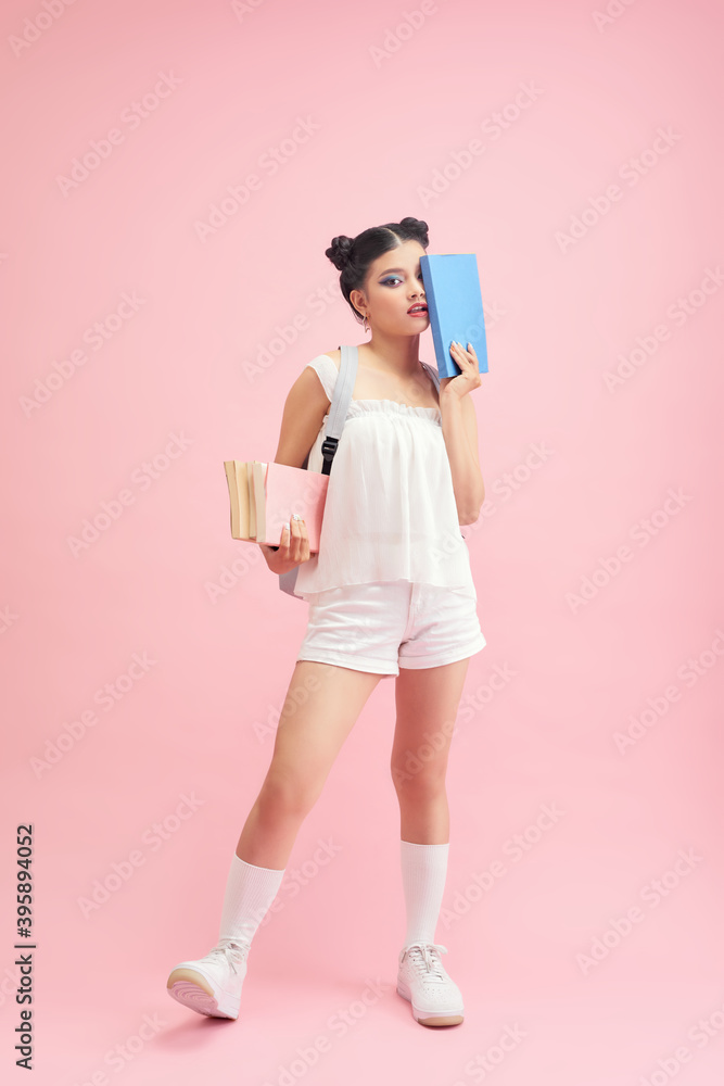 Photo of pretty student girl 20s with double buns hairstyle wearing backpack holding many studying books isolated over pink background Photo of pretty student girl 20s with double buns hairstyle wearing backpack holding many studying books isolated over pink background
