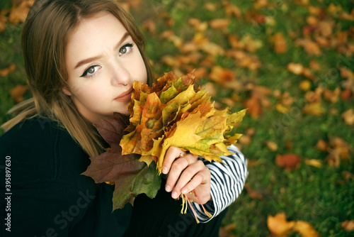 Beautiful young woman sitting on the fallen leaves with a bouquet of maple leaves. cute blond girllooking at camera. top view. photo