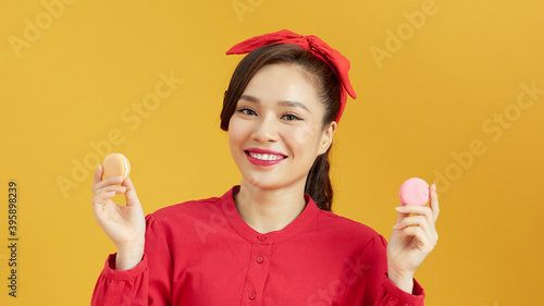 Portrait of an excited woman wearing red dress isolated over yellow background, eating macaroon © makistock