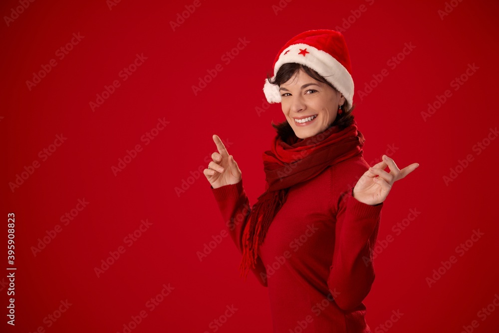 Happy young woman in warm red clothes and santa hat pointing at copy space, looking at camera, smiling. Red background. Happy young woman in warm red clothes and santa hat pointing at copy space, looking at camera, smiling. Red background.