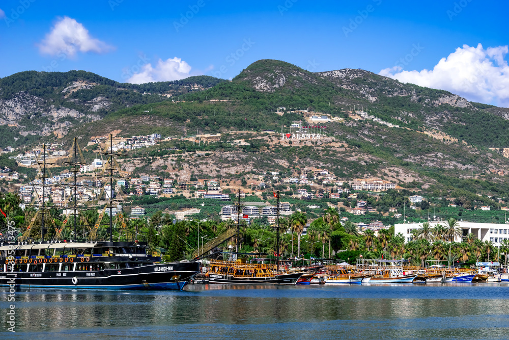 Turkey, Alanya - October 22, 2020: Coastline of Turkish town with sign I Love Alanya on the hill. Beautiful landscape with ships and yachts on the background of a tropical exotic city with palm trees