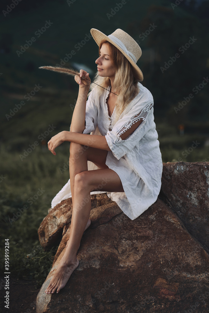 The blonde in a linen dress and straw hat sits on a viewpoint in the mountains. A woman in the Boho style, resting in the countryside, a simple village lifestyle