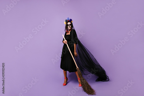 Full-length photo of sorceress with skull mask in black chilling outfit. Woman posing with broom over lilac background