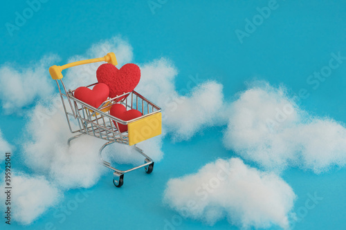 Shopping dream with love. Yellow shopping cart with red hearts inside on a blue background and clouds. Valentine's day.