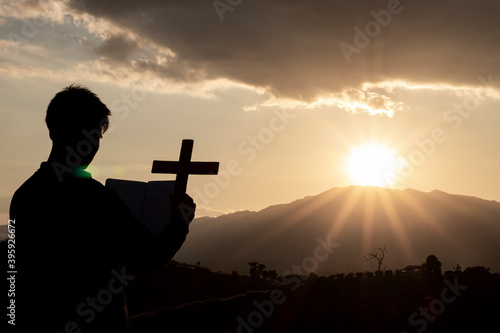 Silhouette of human kneeling down praying and holding christian cross for worshipping God at sunset background. Christian, Christianity, Religion copy space background.