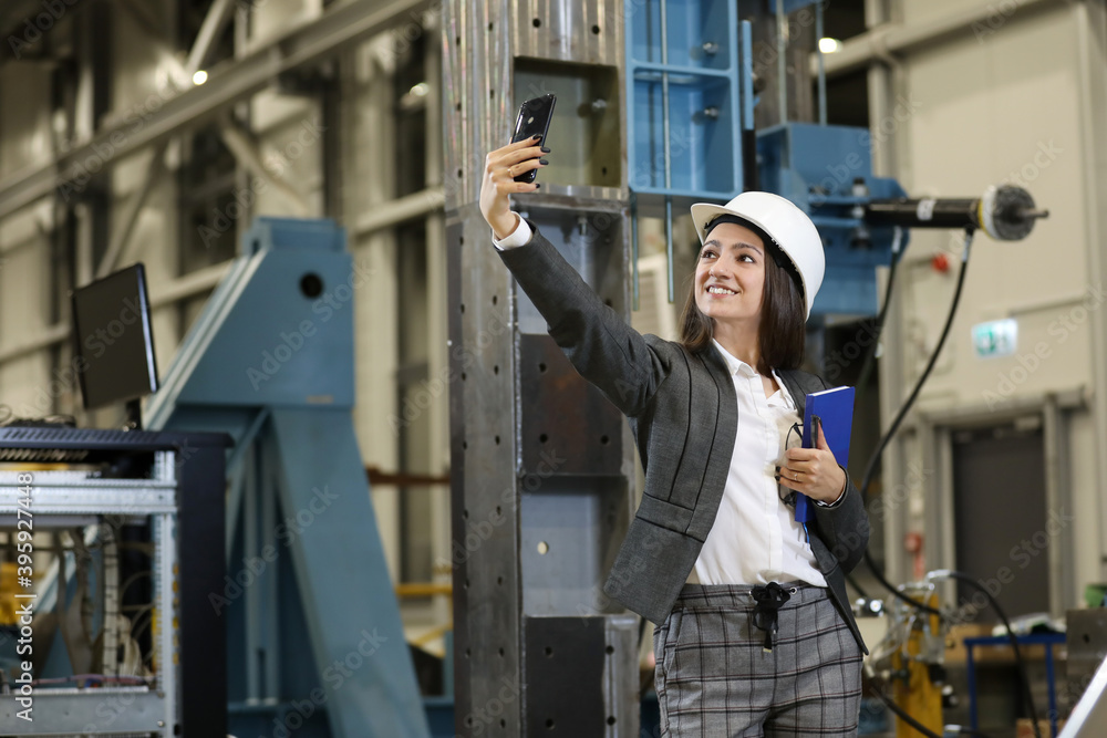 Portrait of a female factory manager in a business suit holding smartphone and notebook, controlling the work process and making selfy in the manufacturer. Portrait of a female factory manager in a business suit holding smartphone and notebook, controlling the work process and making selfy in the manufacturer.