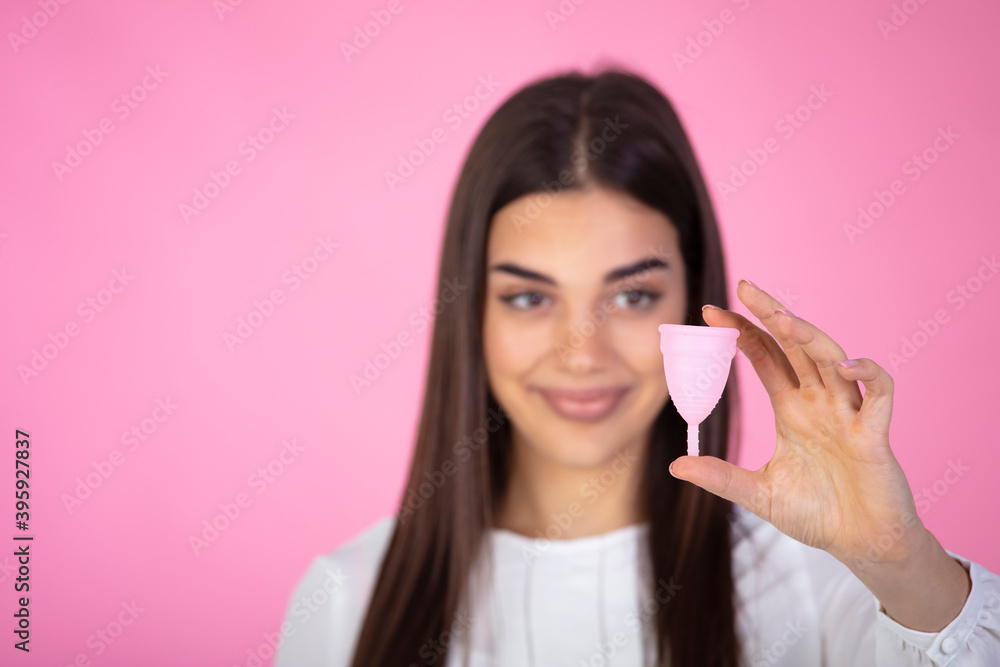 Attractive woman holding menstrual cup standing against pink background, squeesing her shoulders and spreading hand aside, looks smiling at camera. Female period concept. Attractive woman holding menstrual cup standing against pink background, squeesing her shoulders and spreading hand aside, looks smiling at camera. Female period concept.