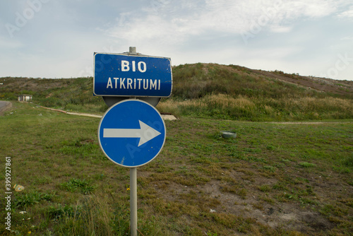 Blue road sign indicating direction. Latvian inscription "BIO waste". Green hill. Cloudy sky.