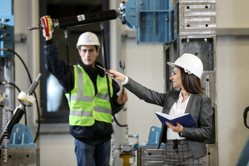 Portrait of a female factory manager in a white hard hat and business suit and factory engineer in work clothes. Controlling the work process in the manufacture. Portrait of a female factory manager in a white hard hat and business suit and factory engineer in work clothes. Controlling the work process in the manufacture.