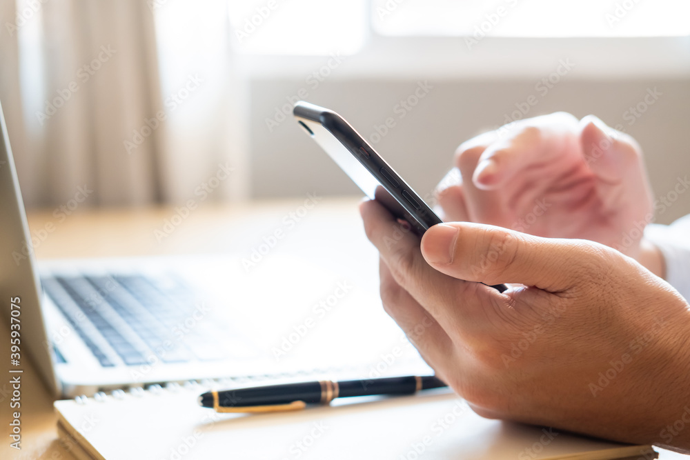 Selective focus at Smartphone. Men hand holding mobile phone while using finger to touch device screen. With blurred computer laptop and paper notebook with pen on working desk. Wireless technology. Selective focus at Smartphone. Men hand holding mobile phone while using finger to touch device screen. With blurred computer laptop and paper notebook with pen on working desk. Wireless technology.
