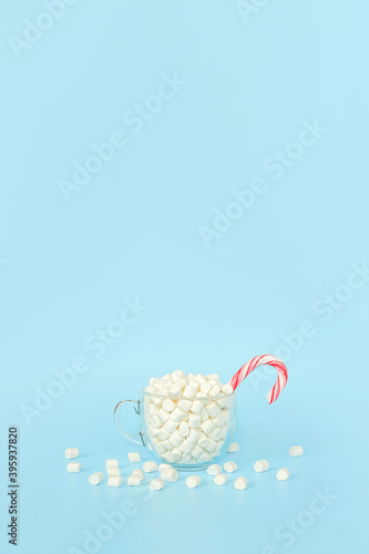 Enjoy winter time, postcard. Big transparent cup of marshmallows with red lollipop cane on blue background. Holiday concept. Front view, Greeting card Copy space