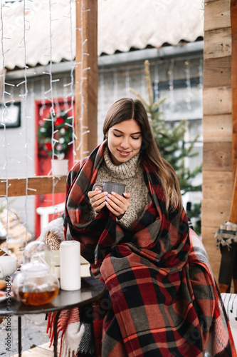 Young caucasian woman in a plaid is drinking a hot drink on the veranda with Christmas tree and decorations.