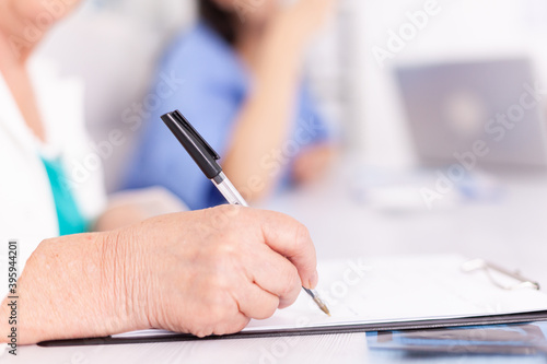 Mature woman doctor taking notes on clipboard during briefing with coworkers. Clinic expert therapist talking with colleagues about disease, medicine professional