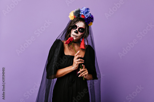 Mysterious Mexican woman in dress of black widow posing on lilac background. Photo of girl with crown of flowers and bright earrings