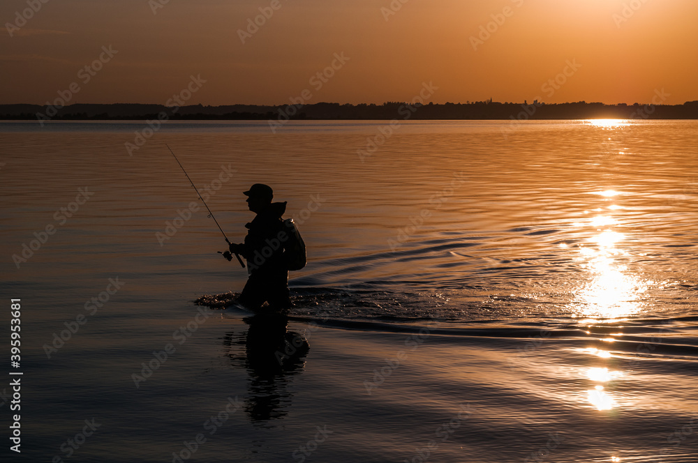 Angler with fishing rod in hand walking in water during sunset in Rewa - Poland.