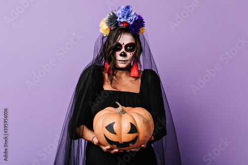 Brunette with face art of Mexican skull holds decorated pumpkin. Photo indoors of young girl with crown of flowers