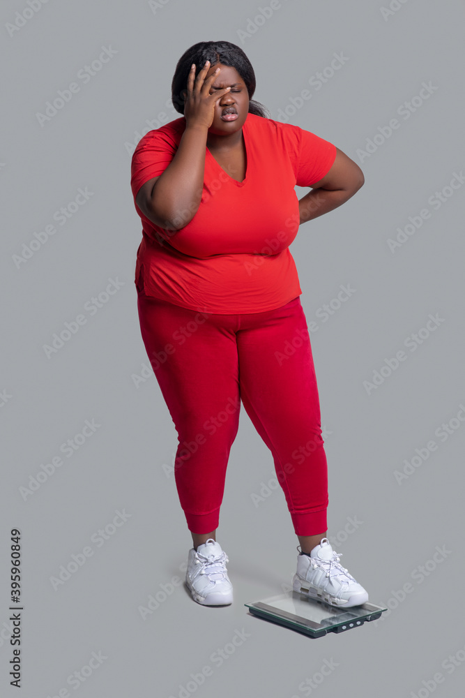 Young dark-skinned woman in red clothes checking weight and feeling disappointed Young dark-skinned woman in red clothes checking weight and feeling disappointed