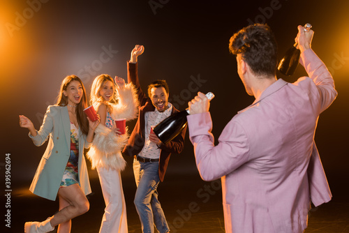 back view of man holding bottles with champagne near excited multicultural friends on black, stock image