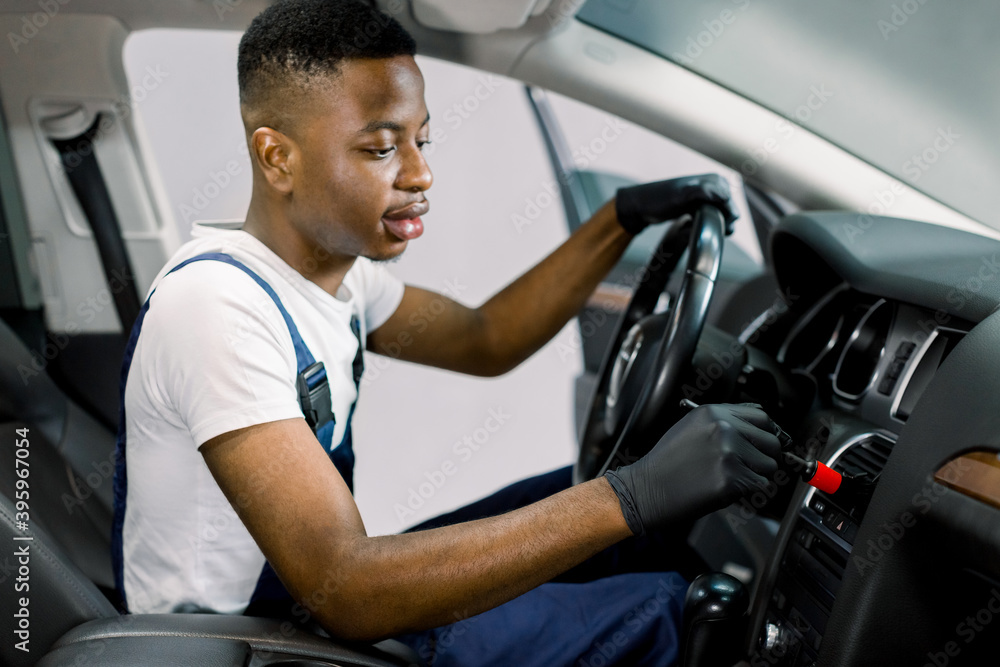 Auto service and professional detailing concept. Young black man worker in white t-shirt and overalls, makes vehicle interior cleaning, wiping front plastic panel with a brush