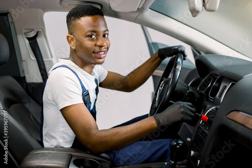 Auto service and professional detailing concept. Young black man worker in white t-shirt and overalls, makes vehicle interior cleaning, wiping front plastic panel with a brush, smiling to camera