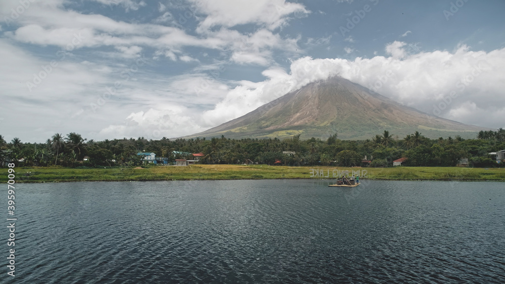 Tropic volcano at lake coast aerial. Tourist cruise at boat at green grass valley at shore. Countryside cottages at palm trees. Nobody nature landscape of Philippines mount Mayon at Legazpi town
