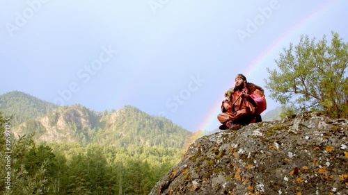 Man in Altai dressed in national clothes sits on rock next to river and plays stringed musical instrument topshur.  Background of mountains and colorful rainbow. Slow motion. Smooth camera movement