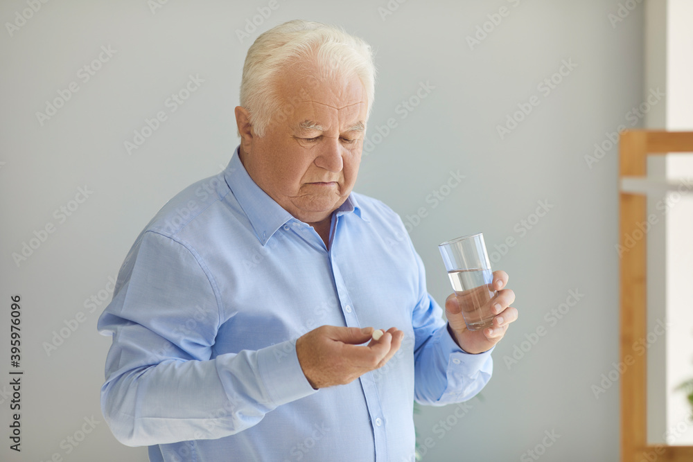 Senior man holding medical pill and glass of water doubting whether the medication is worth it Senior man holding medical pill and glass of water doubting whether the medication is worth it