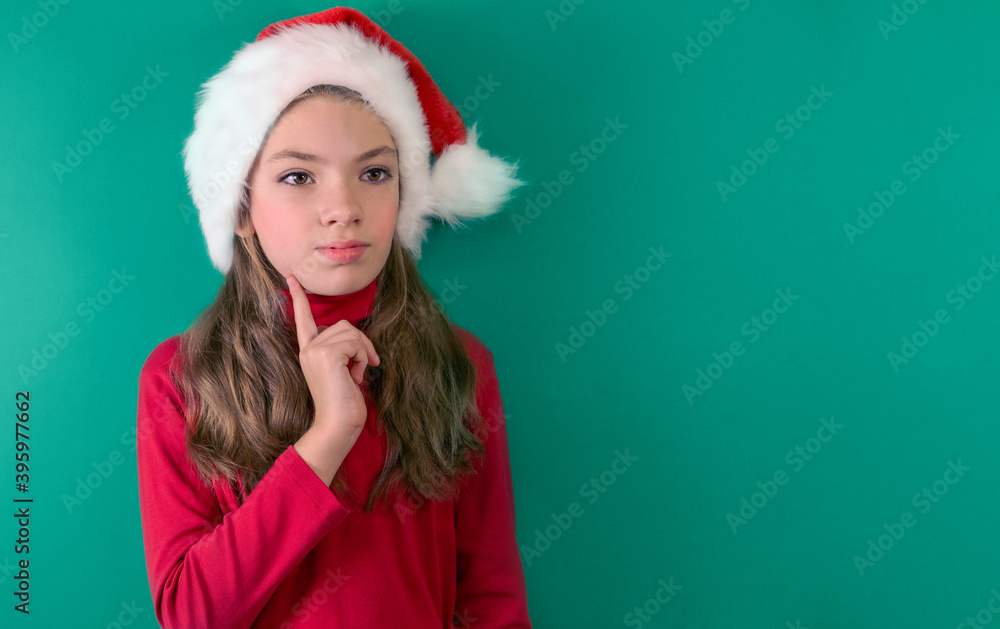 Happy Christmas girl in a red suit and a Santa Claus hat on a green background. Red and green. Portrait of child. Thoughtful look. Space for copying. Happy Christmas girl in a red suit and a Santa Claus hat on a green background. Red and green. Portrait of child. Thoughtful look. Space for copying.