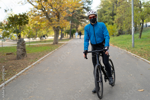An athlete in glasses and a protective mask sits on a bicycle in a park on a bicycle road. Quarantined sports and outdoor recreation.