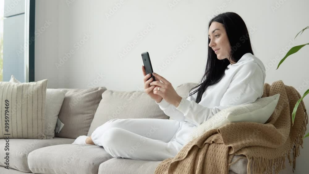 Young woman use phone while sitting on a sofa in a cozy living room, reading books. Stay home concept Young woman use phone while sitting on a sofa in a cozy living room, reading books. Stay home concept