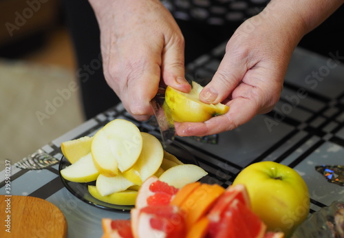 An elderly woman is hands cut an apple into several pieces with a kitchen knife for serving the kitchen table. Healthy homemade food.