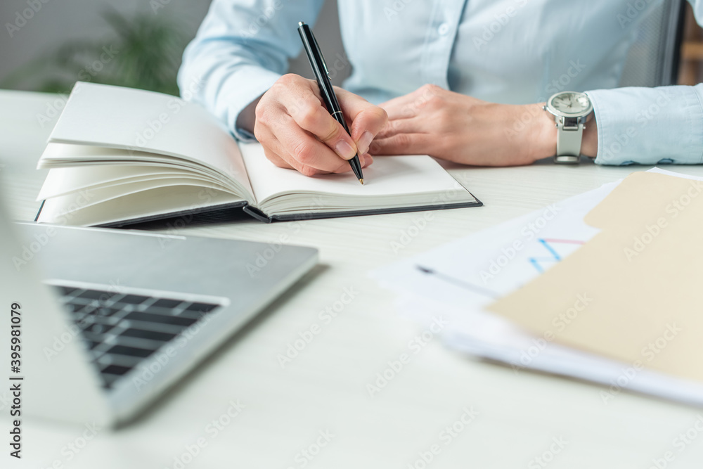 Cropped of businesswoman writing in notebook, while sitting at workplace on blurred foreground Cropped of businesswoman writing in notebook, while sitting at workplace on blurred foreground