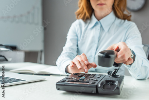 Cropped view of businesswoman with handset dialing number on landline telephone at workplace on blurred background photo