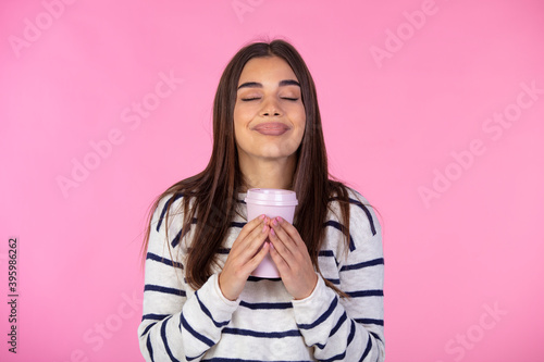 Wonderful aroma. Photo of satisfied woman in sweater holds paper cup of hot drink, closes eyes. Beautiful young woman enjoys aromatic tea or coffee. Isolated on pink background